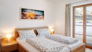 Ein Doppelbett mit floralem Bettbezug, ein Nachttisch mit Lampe, ein Gemälde an der Wand und ein großes Fenster mit Blick auf eine verschneite Landschaft.