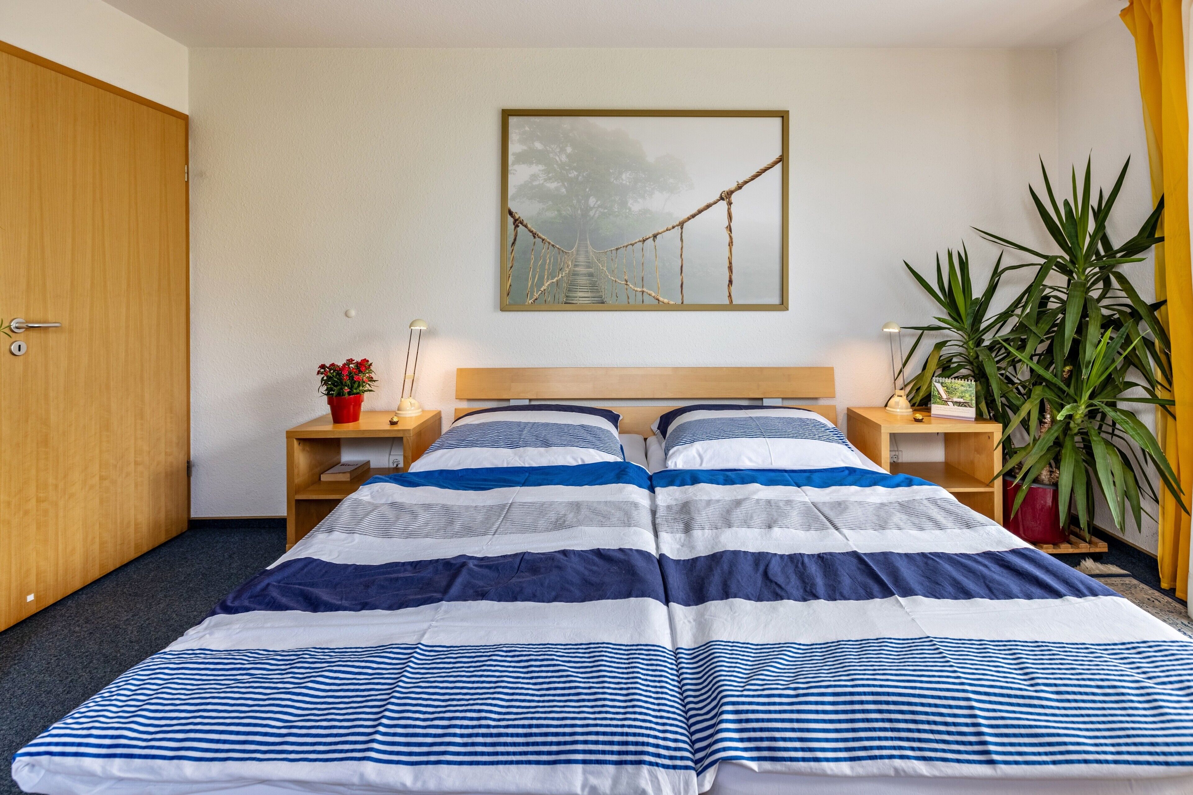 Een tweepersoonsbed met gestreept beddengoed, een houten hoofdbord, nachtkastjes met lampen, een ingelijste foto aan de muur en een potplant.