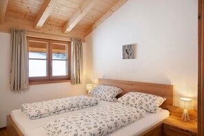 A double bed with floral bedding, a wooden headboard, a nightstand with a lamp, a window with curtains, and a heart-shaped decoration on the wall. - Apartment "Bergenzian" with Mountain View, Balcony & Wi-Fi (Grainau)