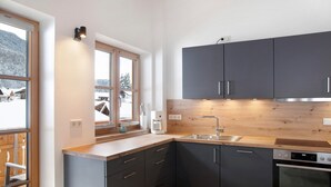 Une cuisine moderne avec des meubles de couleur sombre, un plan de travail en bois, un four encastré et une fenêtre donnant sur un paysage enneigé.