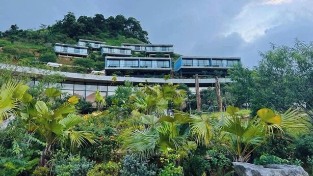 A modern building with multiple levels, surrounded by lush greenery and a hill in the background. - Vigolando Van Ho Resort (Van Ho)