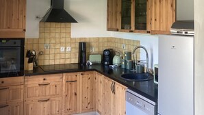 A kitchen with wooden cabinets, a black countertop, a stainless steel refrigerator, and a hanging light fixture. - Villa Morgon (SAVINES LE LAC)