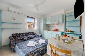 A room with a bed, a dining table with breakfast items, a kitchen area, and a ceiling fan.