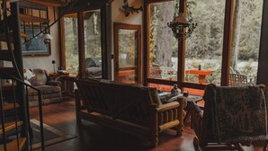 A cozy living room with a wooden staircase, a sofa, a rocking chair, and large windows offering a view of the outdoors.