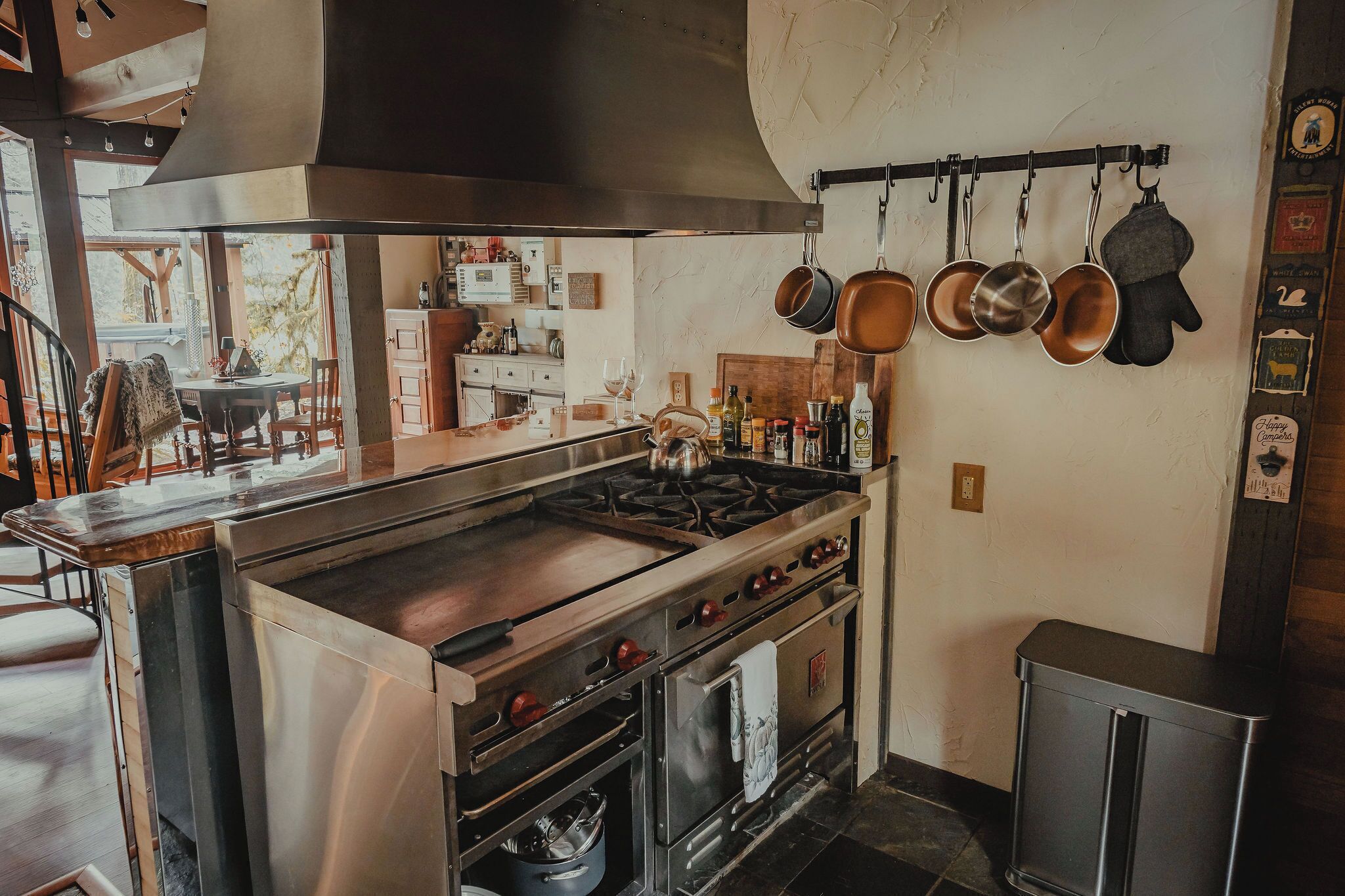 Une cuisine équipée d’une cuisinière, d’un four et de casseroles suspendues. On aperçoit un coin repas avec une table et des chaises à travers une fenêtre.