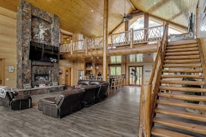 Un intérieur spacieux de chalet en bois, avec une cheminée en pierre, des fauteuils en cuir, un escalier en bois et une grande fenêtre.