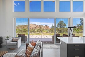 A modern living room with a sofa, armchair, and dining area, featuring large windows and a view of a mountainous landscape.