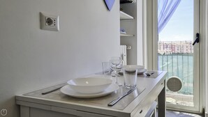 Uma área de refeições com uma mesa posta para dois, uma janela com vista para edifícios e uma tomada na parede.
