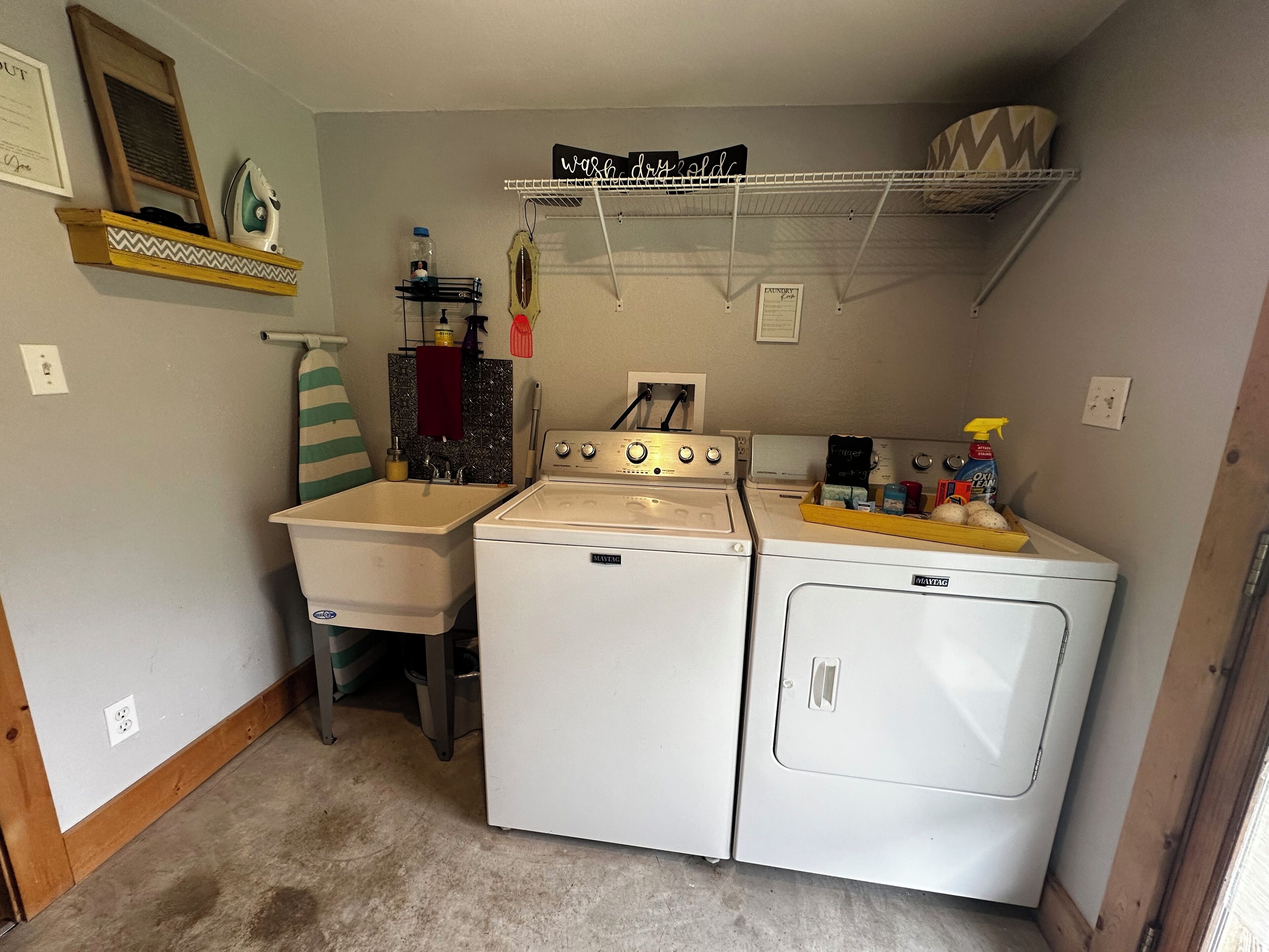 A laundry room with two washing machines, a dryer, and a shelf with laundry supplies.