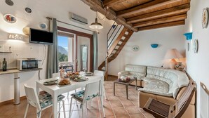 Un interior acogedor con comedor, cocina y zona de estar, que presenta vigas de madera, un televisor y platos decorativos.