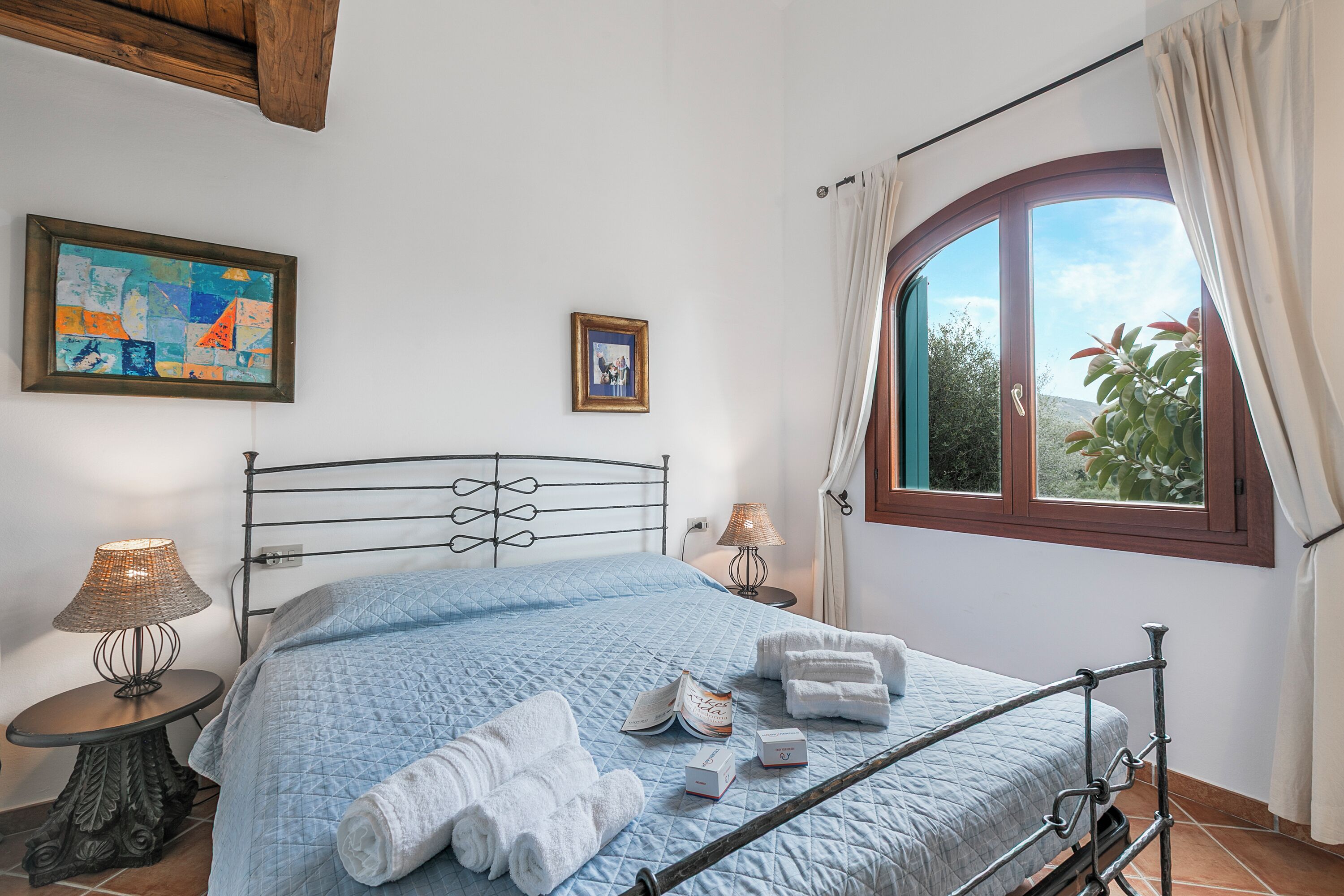 Una cama bien hecha con un edredón azul, dos lámparas de noche y una ventana con cortinas.