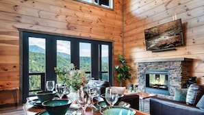 Un interior de madera con una mesa de comedor dispuesta para seis personas, una chimenea y una vista panorámica a través de amplios ventanales.