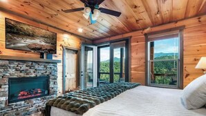 Un dormitorio con chimenea de piedra, cama con manta de piel y ventanas amplias que ofrecen vistas al exterior.