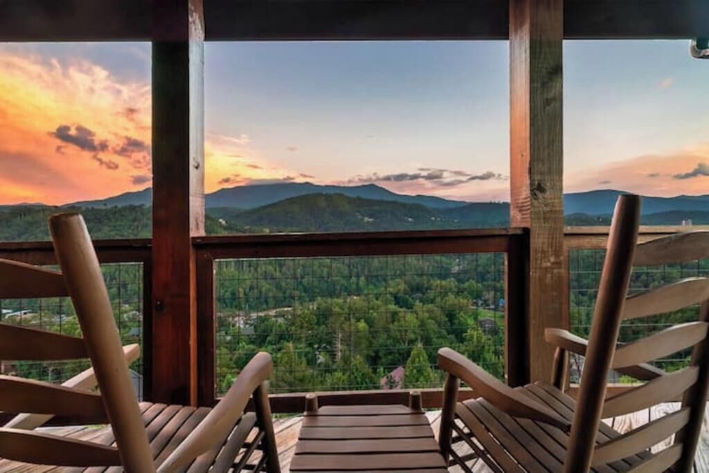 Um deck de madeira com duas cadeiras de madeira voltadas para uma paisagem cênica de montanhas e um pôr do sol.