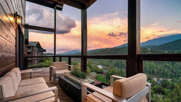 Un área de estar moderna al aire libre con una terraza de madera, sillas con cojines y vistas a un paisaje montañoso al atardecer.