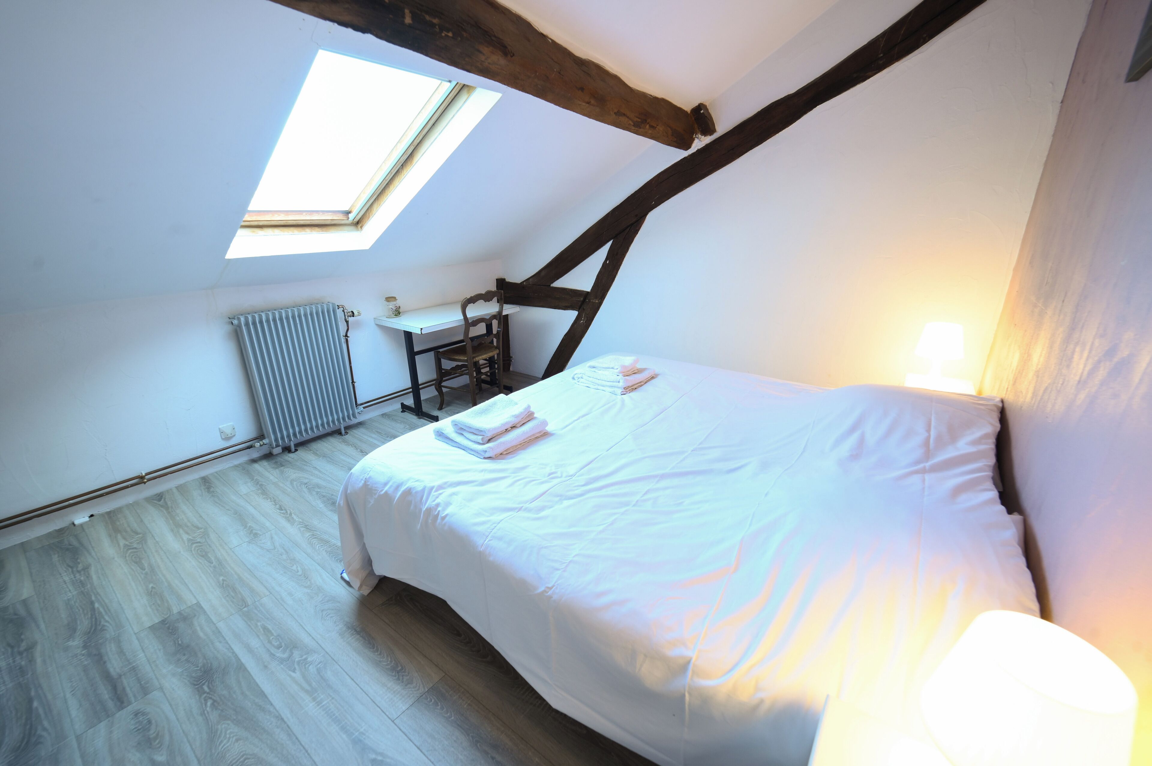 A bedroom with a skylight, a bed with white linens, a radiator, and a small table with a chair.