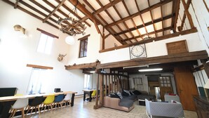 A spacious room with a high ceiling, wooden beams, a chandelier, a clock, and a cozy seating area with cushions.