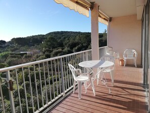 Un balcon avec une table et des chaises, une vue sur une colline boisée et une balustrade blanche.