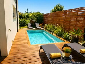 Una piscina rectangular con un deck de madera, sillas de descanso y plantas en macetas.