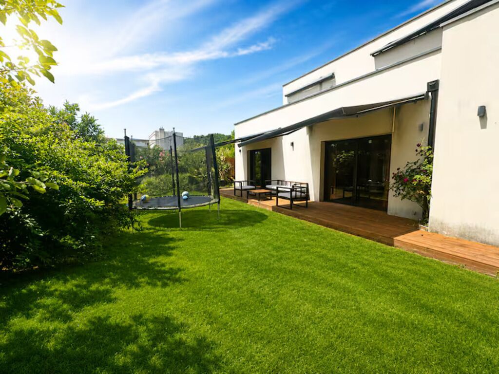 Una casa moderna con un césped cuidado, un patio con mobiliario exterior y una entrada amplia con ventanas.