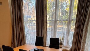 A dining area with a wooden table and black chairs, large windows with curtains, and a view of trees and a street.