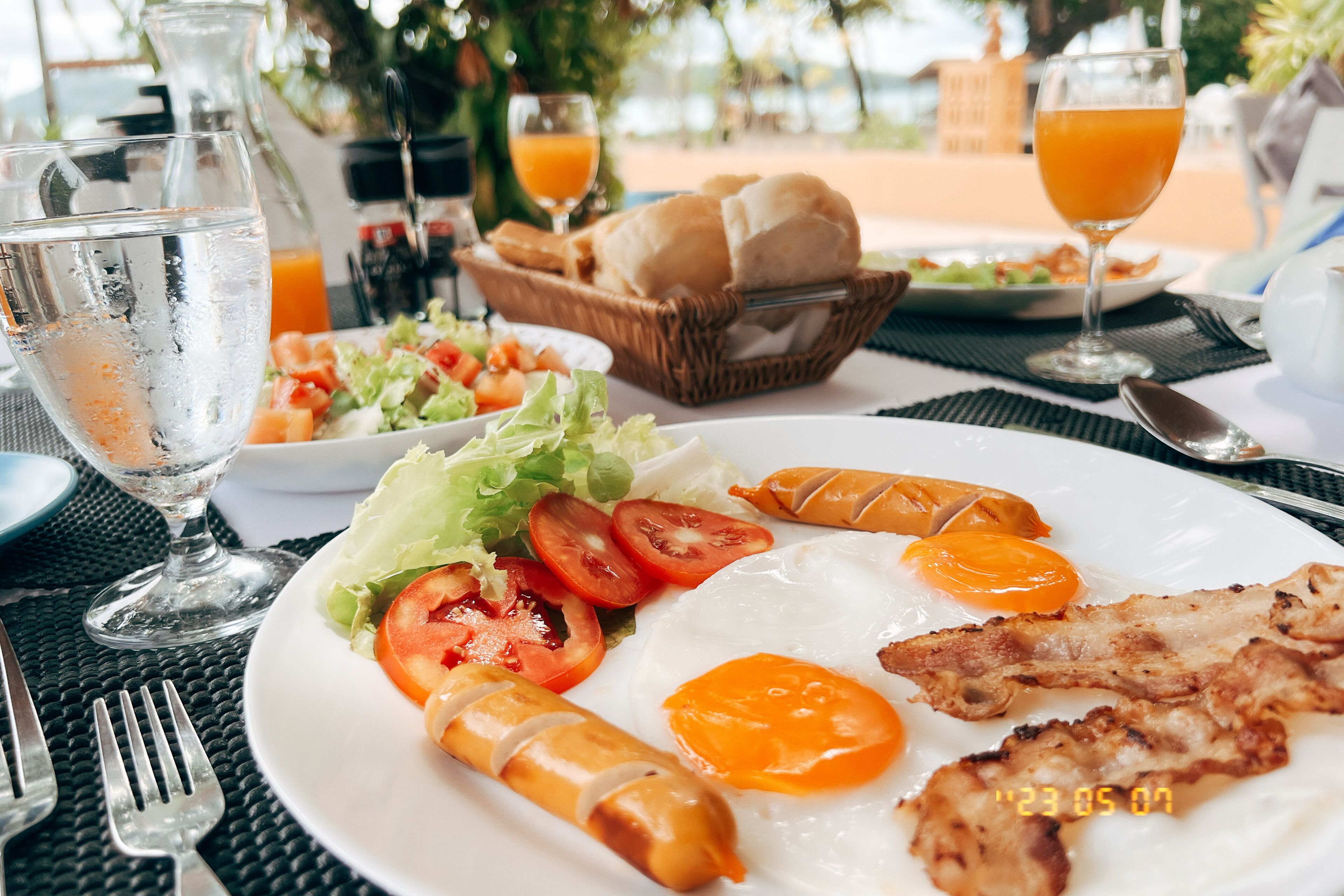 A breakfast setting with a plate of eggs, sausages, and tomatoes, a basket of bread, and a glass of orange juice.