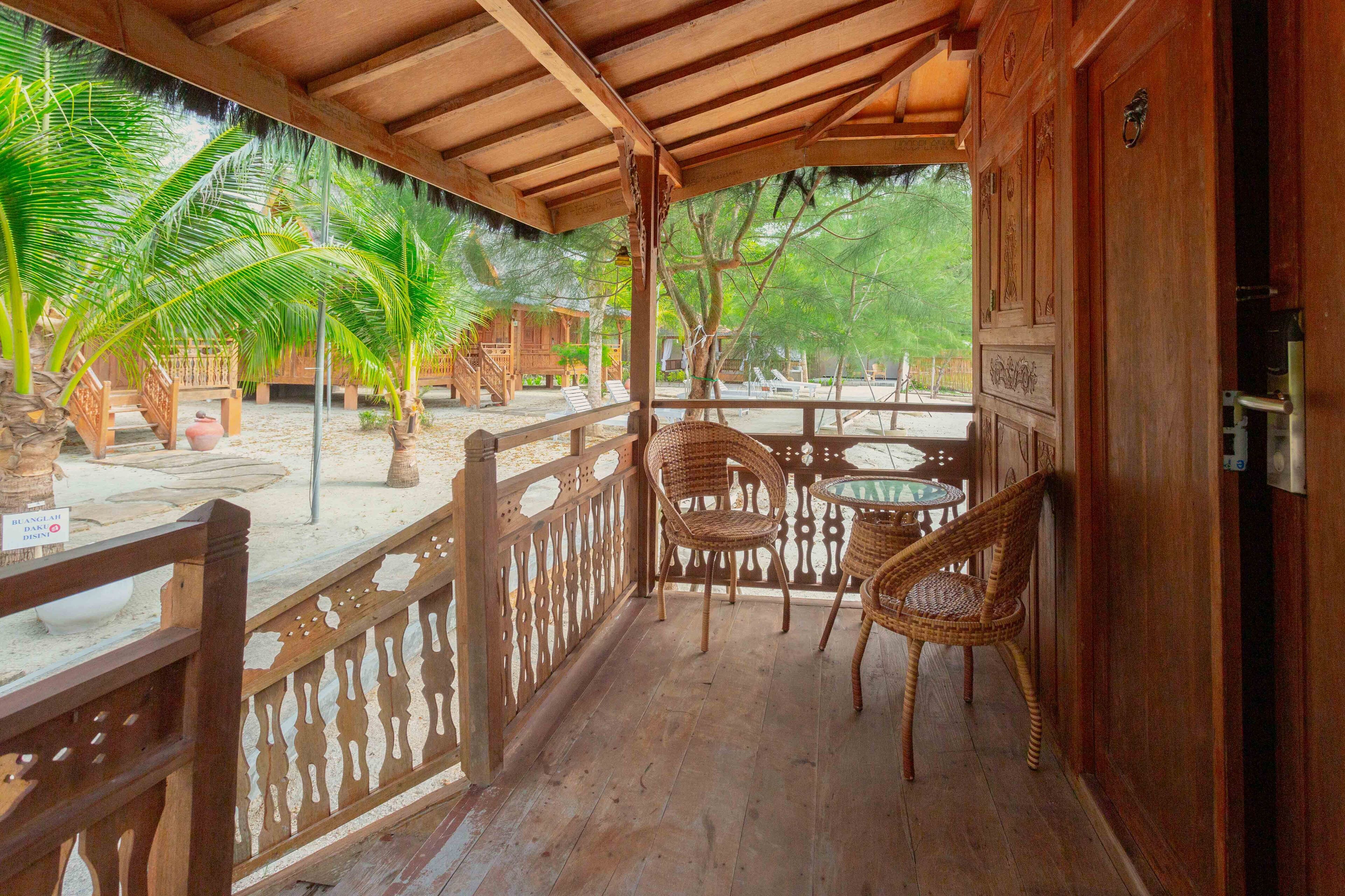 A wooden balcony with wicker chairs and a table, overlooking a sandy area with palm trees.