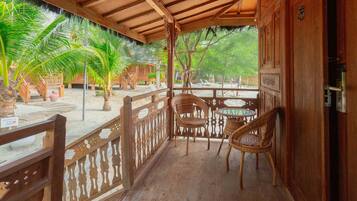 A wooden balcony with wicker chairs and a table, overlooking a sandy area with palm trees.