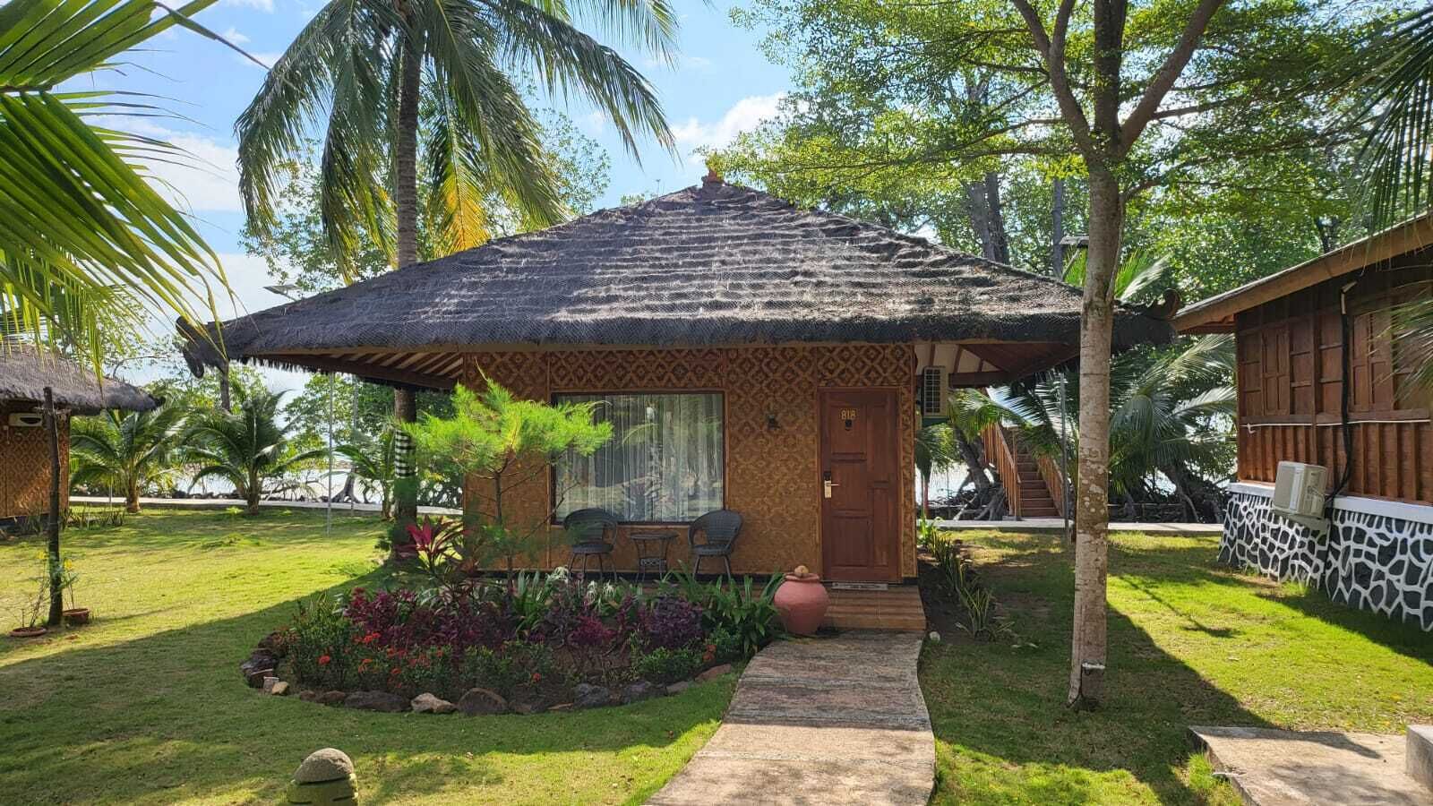 A small, thatched-roof structure with a wooden door, surrounded by a garden with various plants and a palm tree.