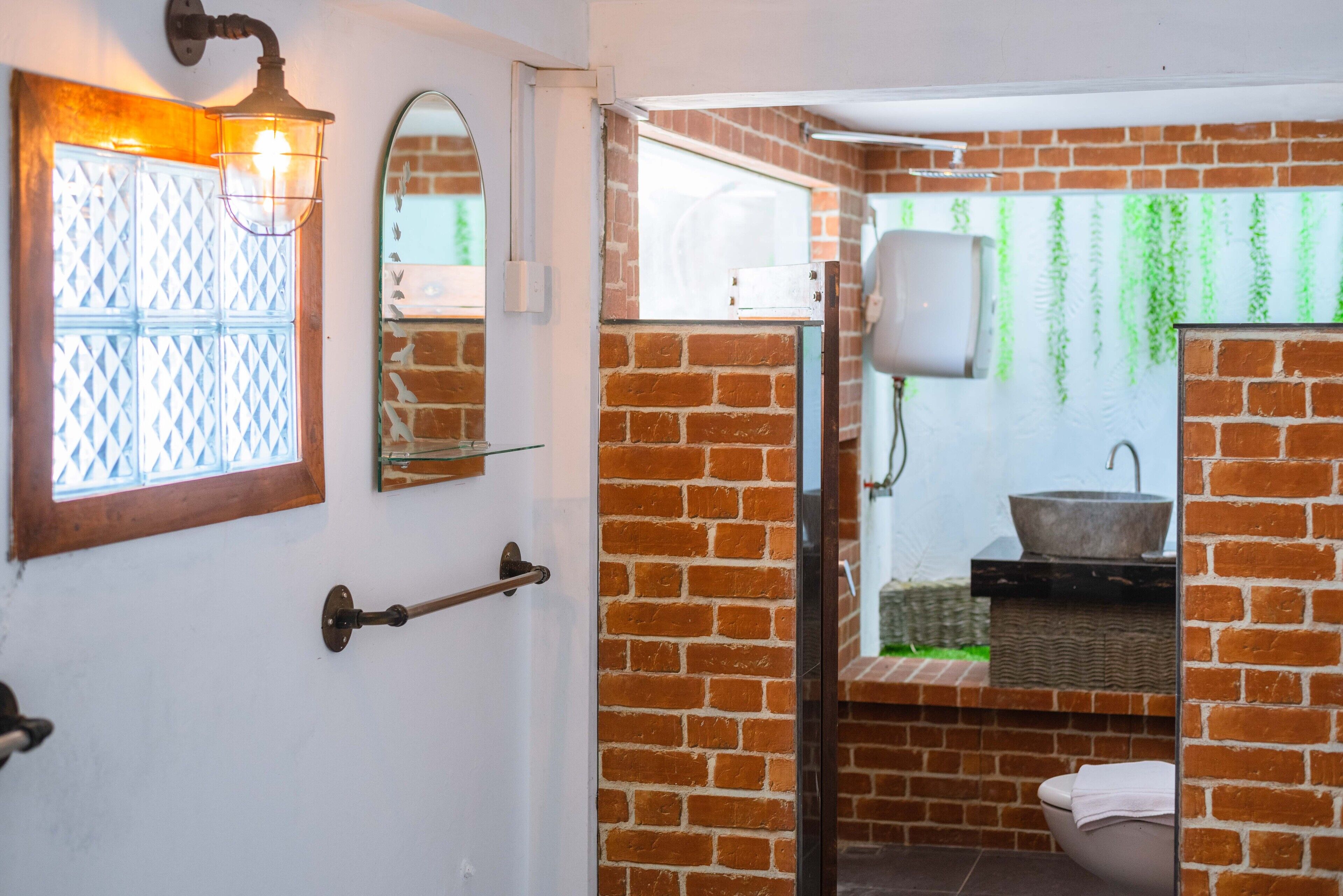 Une salle de bain avec un mur en briques, une porte blanche, une fenêtre avec des vitres à motifs géométriques, un lavabo blanc et des toilettes blanches.