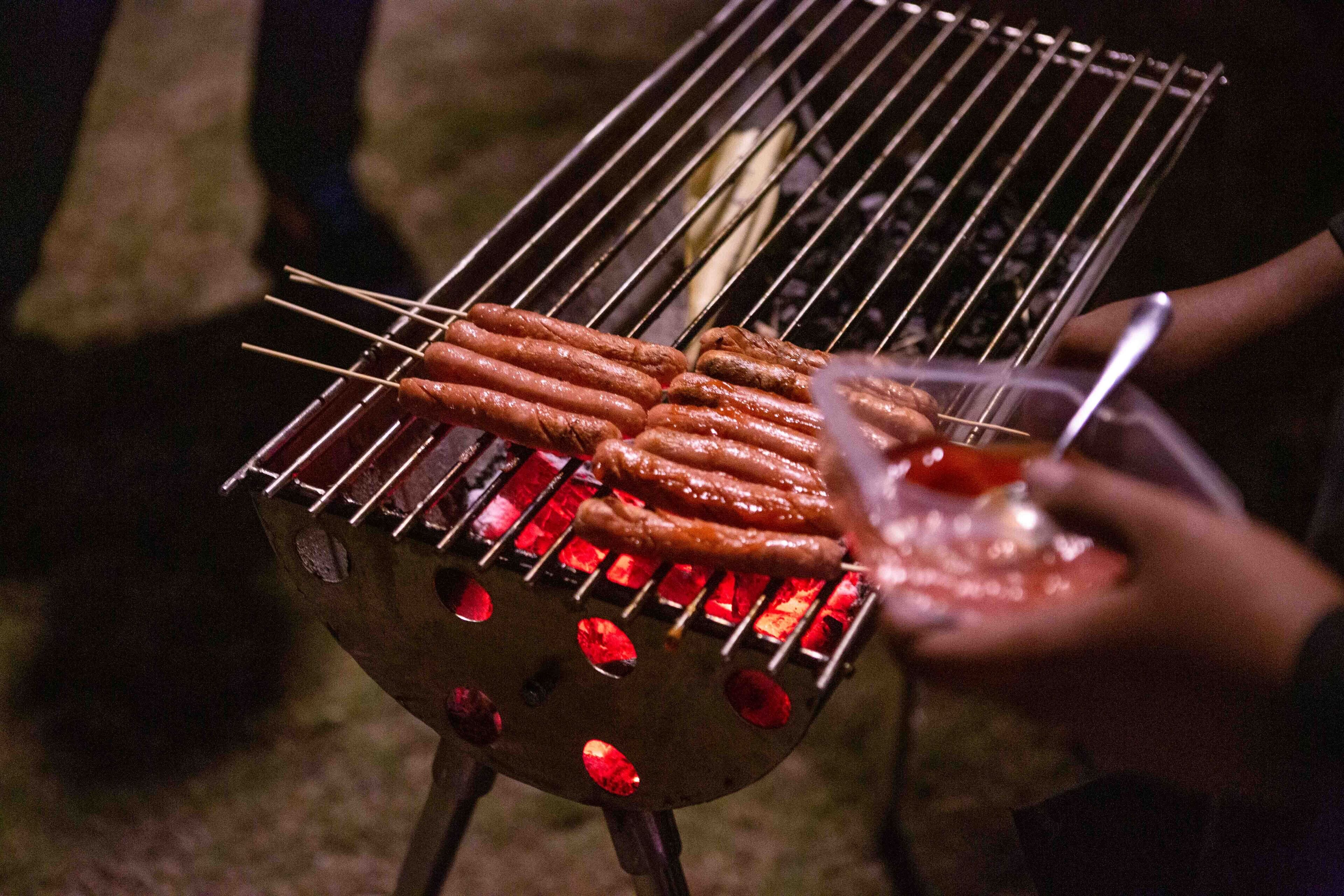 A grill with sausages being cooked, a person holding a skewer, and a glass with a beverage.