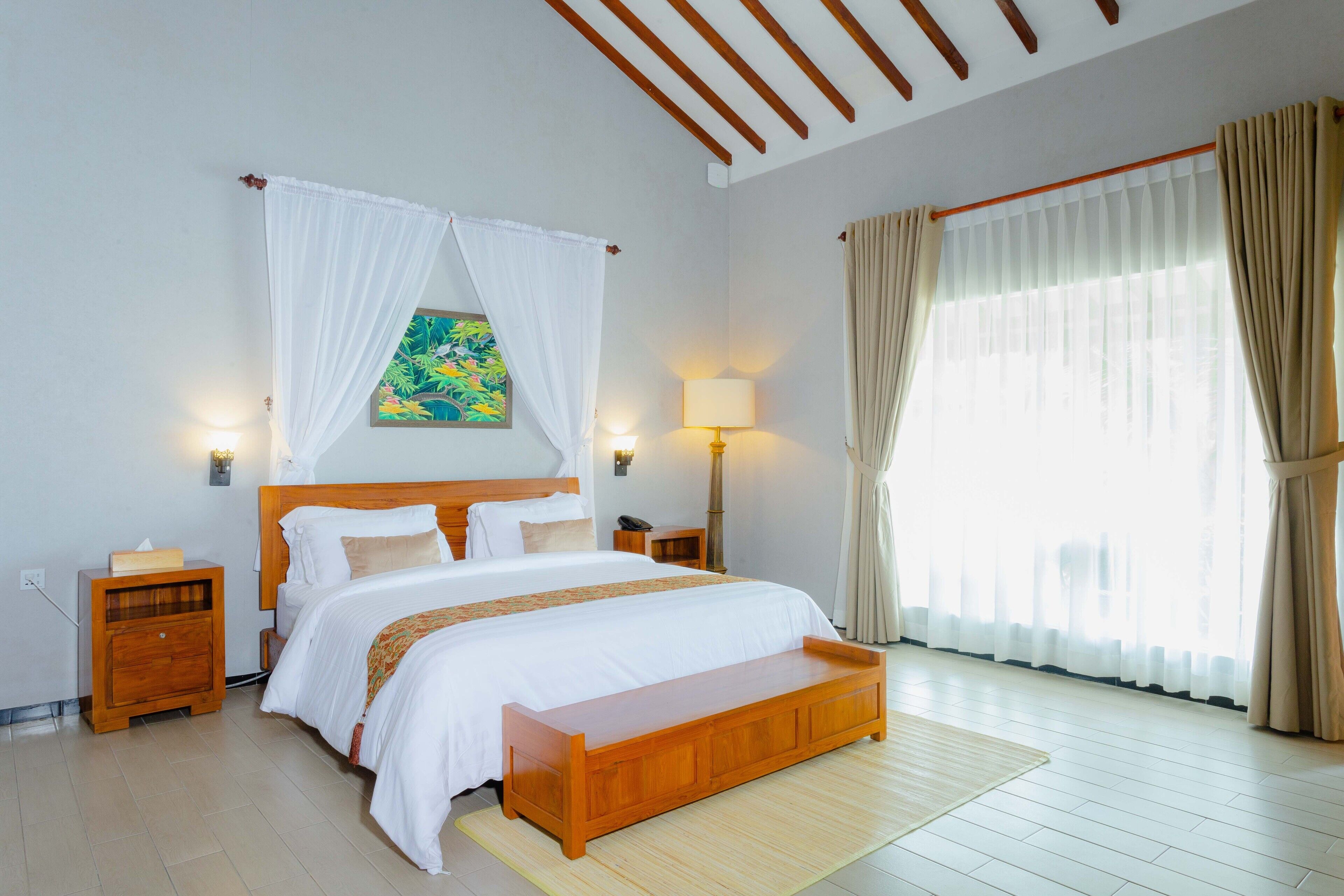 A bedroom with a large bed, wooden headboard, bedside tables, a bench, a painting, and curtains.