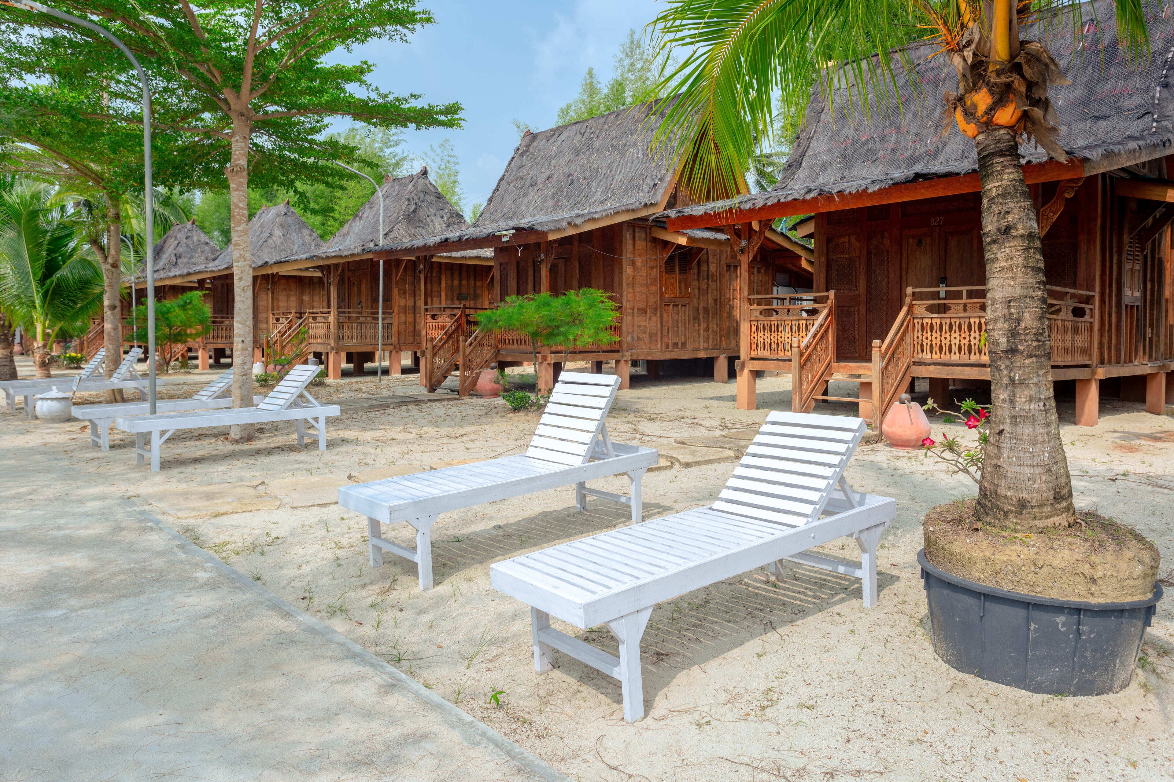 A beachfront resort with wooden huts, white lounge chairs, and a palm tree.