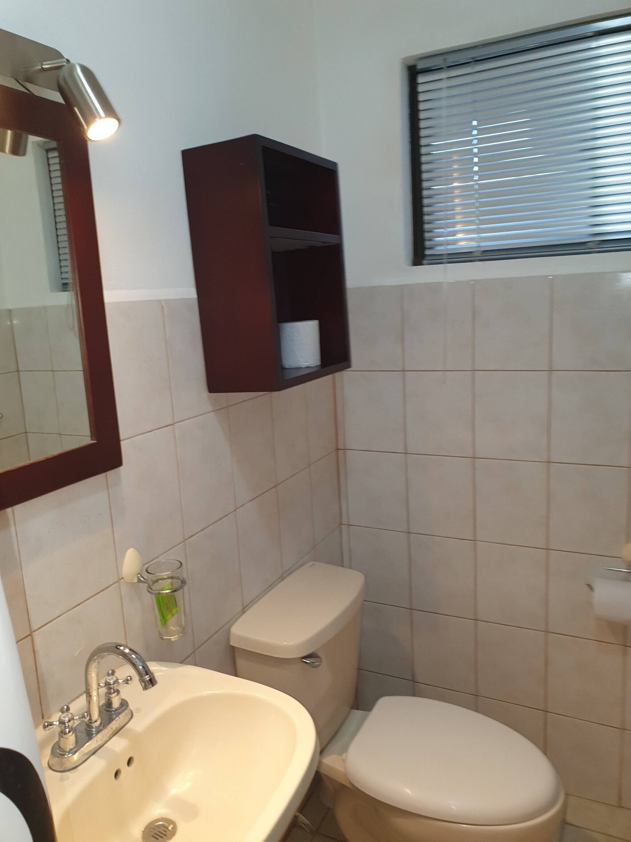 A bathroom with a sink, toilet, mirror, and a shelf above the toilet.