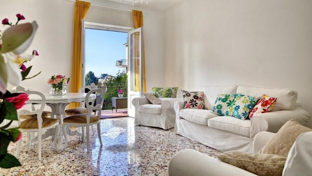A living room with a white sofa, a dining table with chairs, a chandelier, and a view through an open door. - Central apartment Stella House (Massa Lubrense)