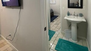 Un baño con un lavamanos blanco, un espejo, un televisor montado en la pared y una puerta que da a otra habitación.