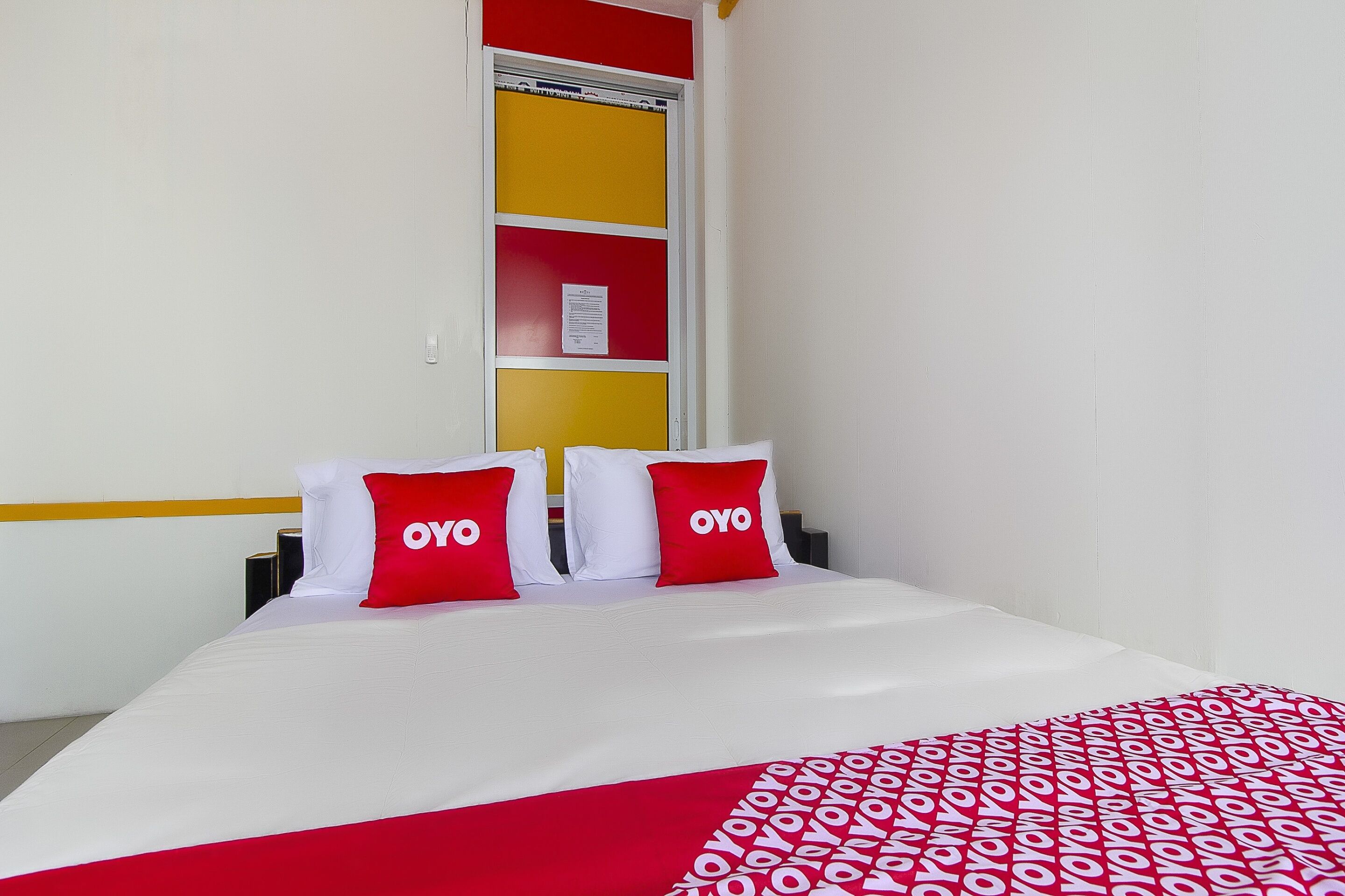 A hotel room with a bed, two red pillows with the word "OYO" on them, a red and white patterned blanket, and a window with a yellow and red wall panel.