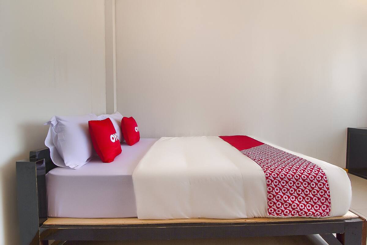 A neatly made bed with white and red bedding, a wooden headboard, and a bedside table with a black object on it.
