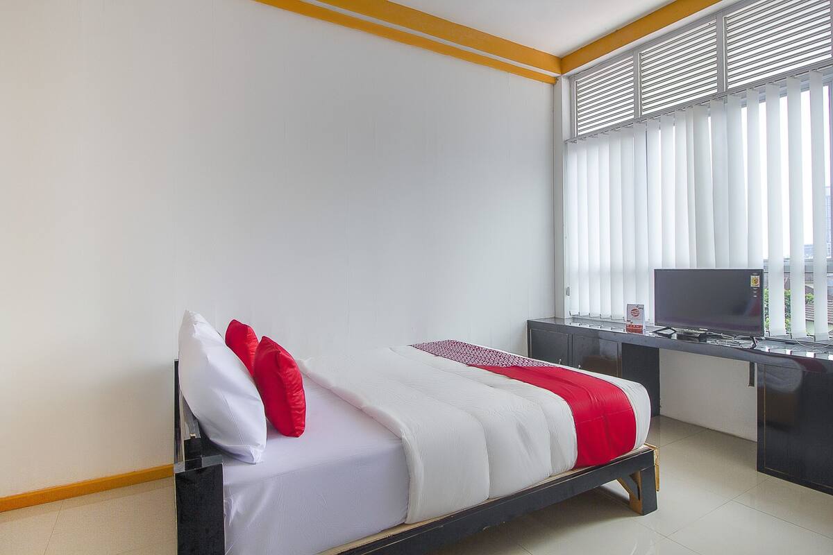 A bed with white and red bedding, a desk with a TV, and a window with blinds.