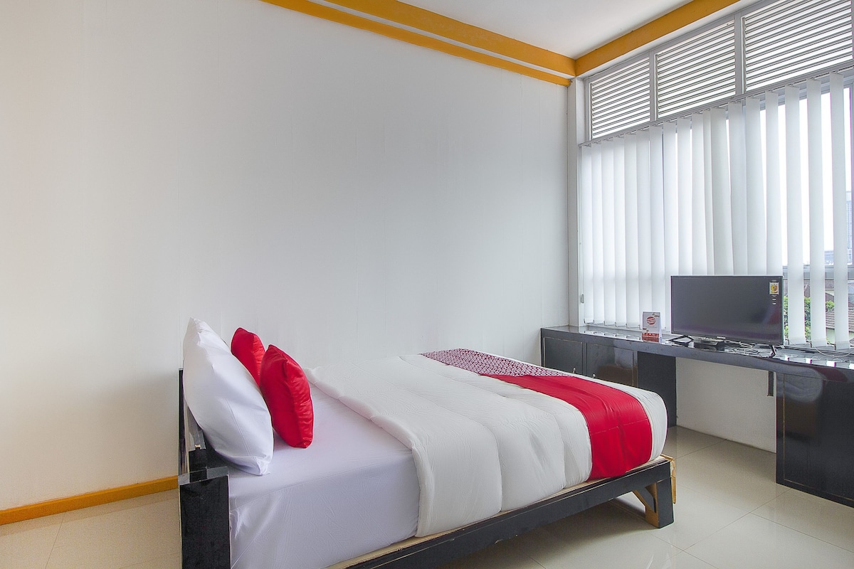 A bed with white and red bedding, a desk with a TV, and a window with blinds.