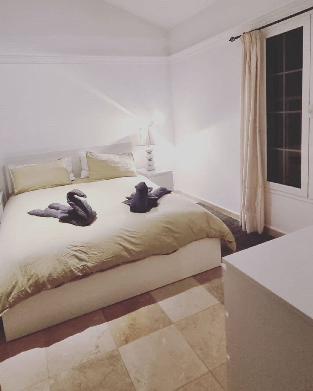 Ein Schlafzimmer mit einem Bett, Kissen, einem Nachttisch, einer Lampe und einem Fenster mit Vorhängen.
