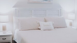 Une chambre à coucher blanche avec un lit, des tables de chevet et un tableau représentant une scène de plage.