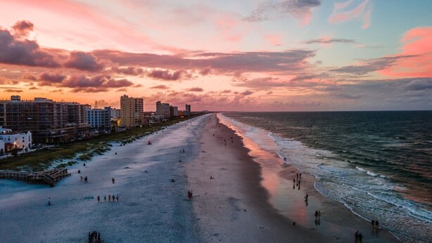 Une plage où des personnes se promènent le long du rivage, avec des bâtiments en arrière-plan et un ciel au coucher du soleil.