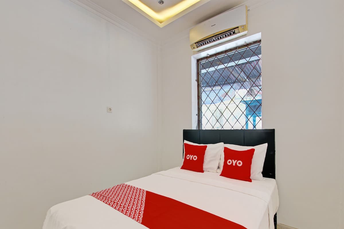 A hotel room with a bed, a window with a metal grill, and an air conditioning unit above the window.