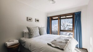 A bedroom with a bed, two pillows, a blanket, a nightstand with a towel, and a window with curtains. - Charming House With all the Home Comforts (Arrowtown)