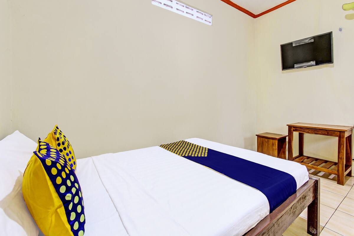A single bed with a white and blue bedspread, a yellow pillow with a polka dot pattern, and a small wooden nightstand with a TV on the wall.
