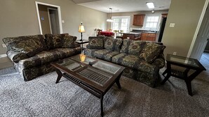 A living room with a sectional sofa, a glass coffee table, and a small side table. There is a kitchen in the background with wooden cabinets and a window. - Life is Good in Mayflower (Mayflower)