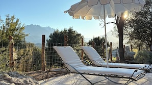 A sunlit outdoor seating area with lounge chairs and a large umbrella, surrounded by a natural landscape and a mountain view.