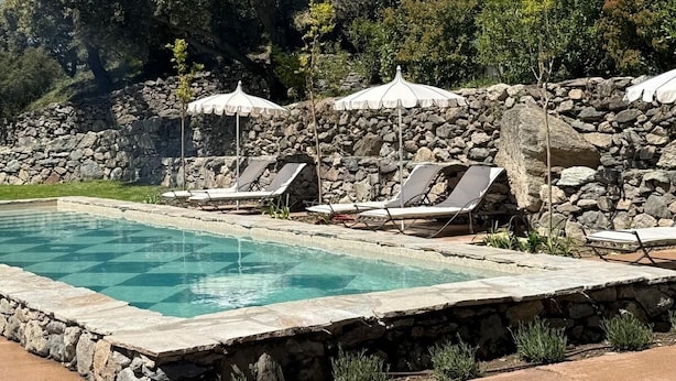 A pool with stone walls, surrounded by lounge chairs and umbrellas, set against a backdrop of trees and a rocky hillside.