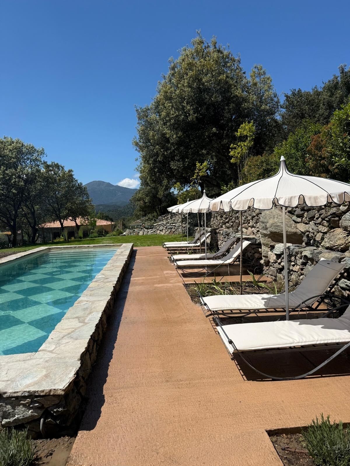 A poolside area with lounge chairs and umbrellas, a stone pathway, and a view of mountains.